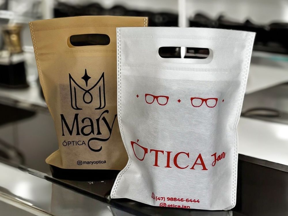 "Sacolas personalizadas de duas óticas, uma dourada da Mary Óptica e outra branca da Ótica Jan, expostas sobre balcão moderno."