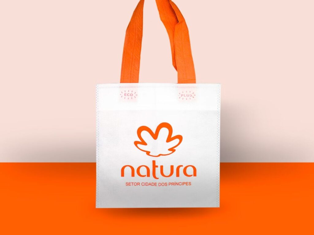 Sacola personalizada da marca Natura com alças laranjas e corpo branco em TNT, reforçando a importância da embalagem como ferramenta de marketing sustentável.