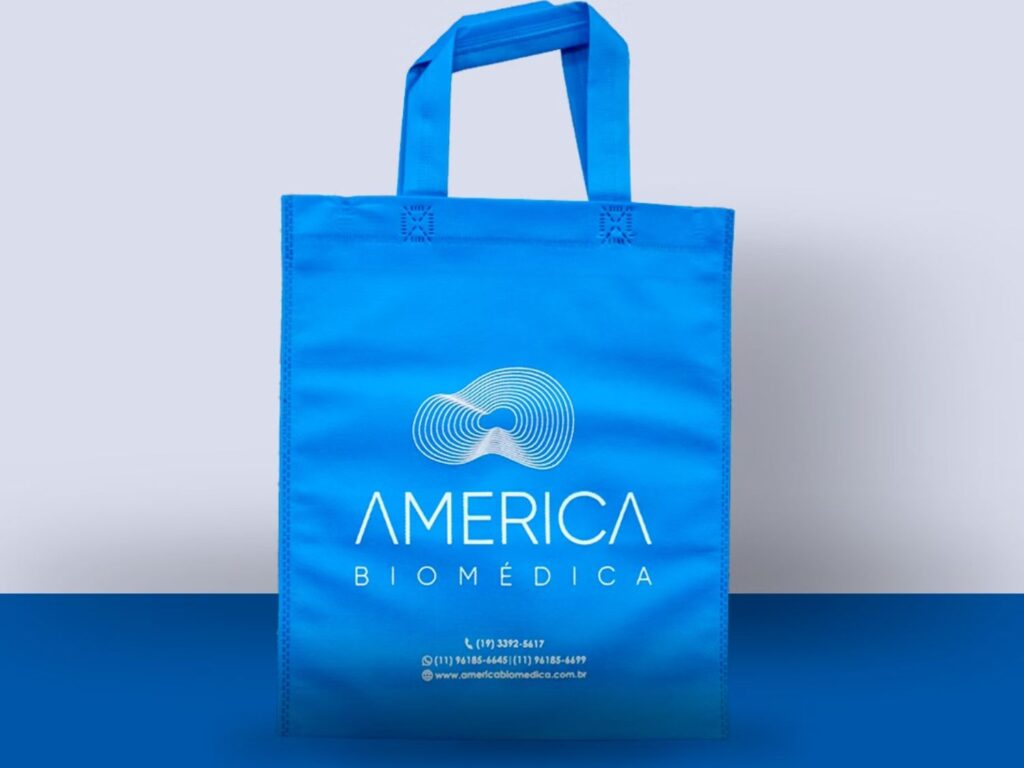 Sacola azul em TNT com alças longas e estampa da América Biomédica, representando a importância da embalagem ecológica na identidade visual da marca.