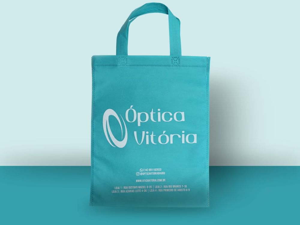 Sacola azul de tecido com alça, personalizada com a logo da Óptica Vitória e informações de contato.