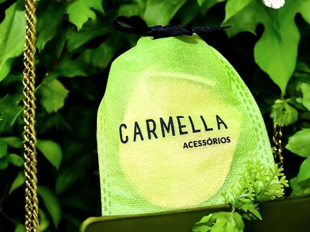 Sacola de tecido verde personalizada com a marca Carmella Acessórios, usada como lembrancinha elegante entre folhagens decorativas.