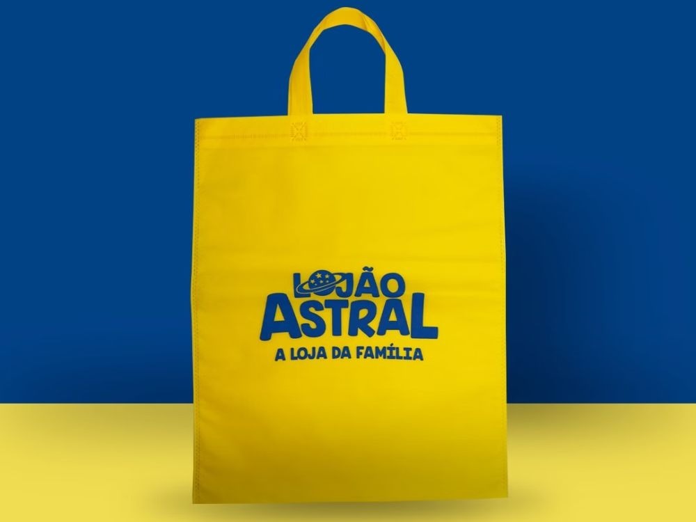 Sacola retornável amarela personalizada da João Astral em fundo azul, apresentada como opção de Brindes úteis e criativos para ações promocionais.