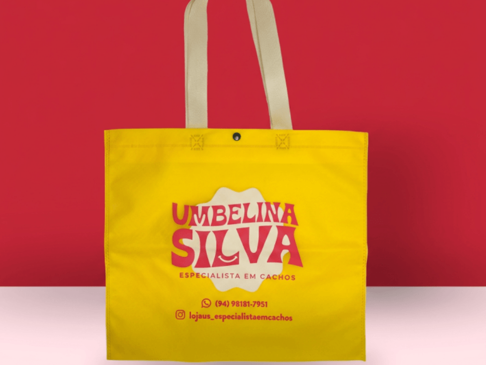 Sacola de tecido TNT amarela com alças longas e personalização em vermelho, exibida sobre fundo vermelho e claro.