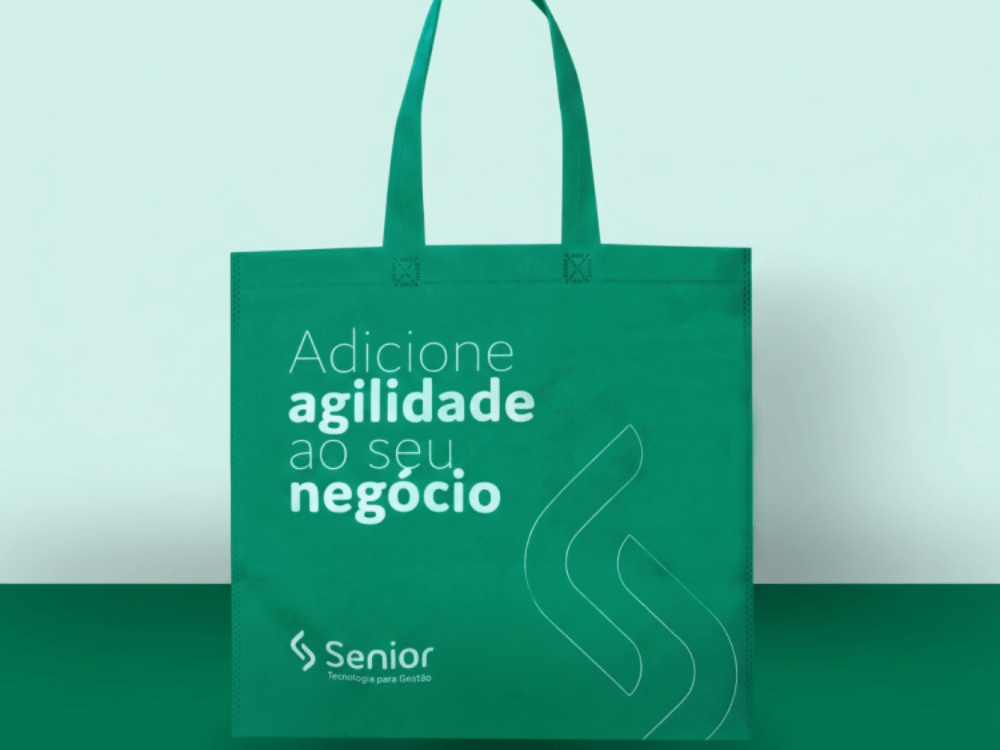 Sacola de tecido TNT verde com alças longas e mensagem personalizada, posicionada diante de um fundo em tons de verde.