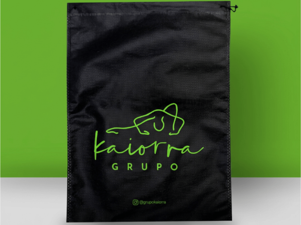 Sacola de TNT preta personalizada com logotipo verde e cordão para fechamento, apresentada em fundo verde e branco.
