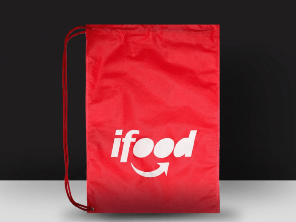 Sacola de TNT vermelha com fechamento por cordão e logotipo branco da iFood, destacada sobre fundo escuro.