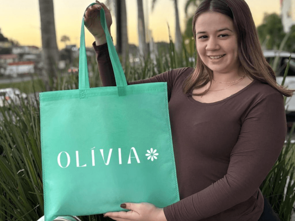 Mulher sorrindo ao segurar uma sacola verde personalizada com alças e logotipo Olivia em área externa arborizada.