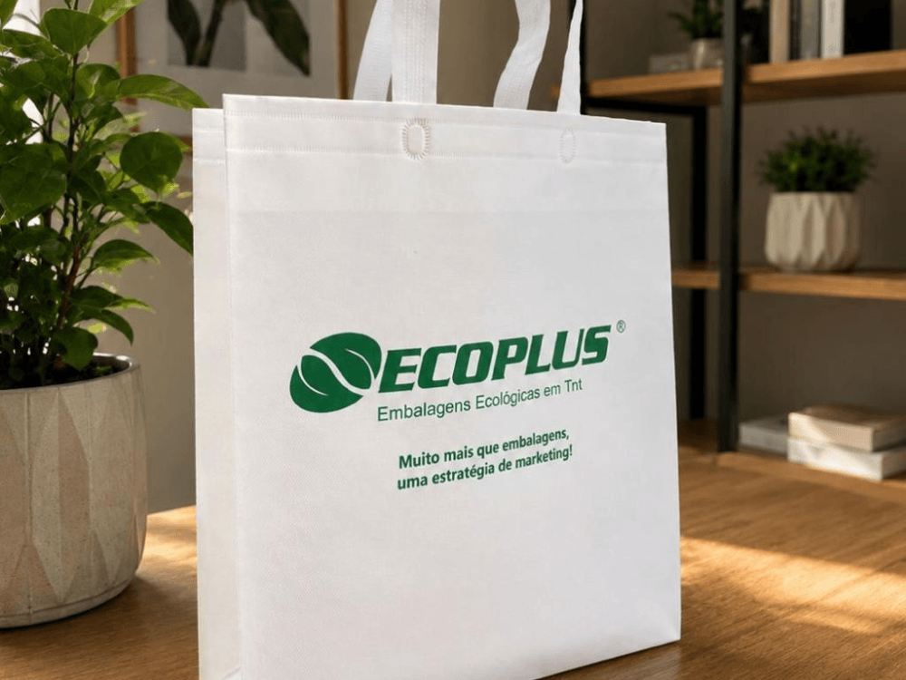 Sacola branca de TNT com alças e logotipo Ecoplus sobre mesa de madeira, em ambiente interno com plantas decorativas.